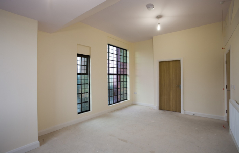 A light empty room with a wood door and two different sized windows with multicoloured panes of glass.