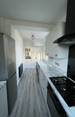 A section of a light narrow kitchen showing the oven and hobs, fridge and other kitceh essentials.